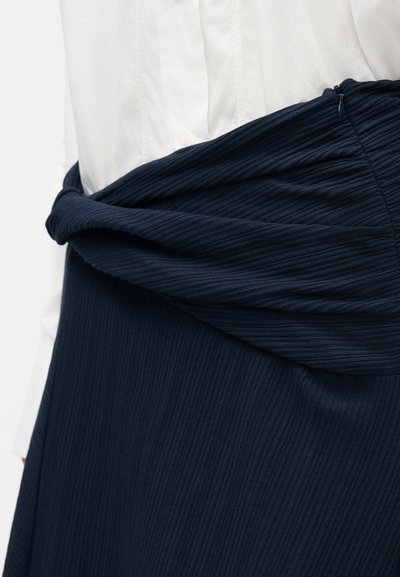 Κοντινό πλάνο ενός ατόμου που φοράει λευκό πουκάμισο και φούστα από μπλε ναυτικό ριμπ ύφασμα με στριφτή ζώνη στη μέση και πλευρικό φερμουάρ.