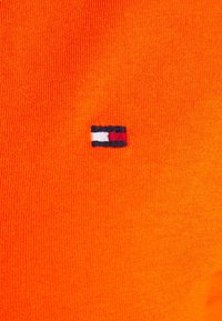 Lys orange strikket stof med et lille broderet logo i marineblå, hvid og rød i nederste venstre hjørne. Glat tekstur, afslappet design.