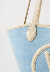 Gaiši zila denim soma ar krēmkrāsas ādas akcentiem, ar noapaļotu pamatni un izteiktu logotipa detaļu sānos.