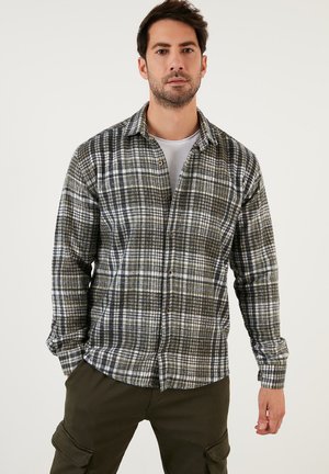 Camisa de franela con un patrón de cuadros en verde, negro y blanco, que presenta un cuello, cierre de botones en el frente y puños. La tela parece suave y texturada.