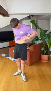 Fiatal férfi lila ingben és fekete rövidnadrágban egy téglalap alakú eszközt helyez a derekán lévő zsebbe beltéren.