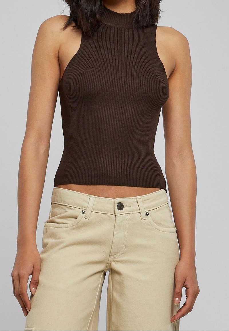 Femme portant un haut sans manches côtelé de couleur marron foncé et un pantalon beige clair, debout devant un fond gris uni.