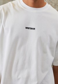 T-shirt en coton blanc avec col rond et manches courtes. L'avant présente le texte noir brodé "WRSTBHVR" centré sur la poitrine.