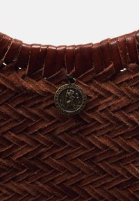 Hnedá tkaná kožená taška s textúrovaným dizajnom a pripevneným okrúhlym príveskom so svätým Kristiánom pre detaily.