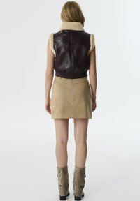Chaleco sin mangas de cuero marrón con cuello de borreguillo crema; combinado con una mini falda de ante beige. Botas altas de ante gris con botones decorativos.