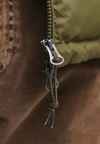 Giacca verde oliva con zip, dotata di una doppia cerniera in metallo e texture in tessuto marrone sullo sfondo; presenta dettagli in paracord sul tira-zip.