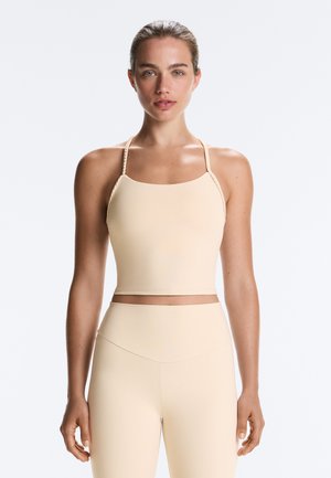 Kvinde står med fronten mod kameraet iført en beige ærmeløs cropped top med tynde flettede stropper og matchende bukser med høj talje.