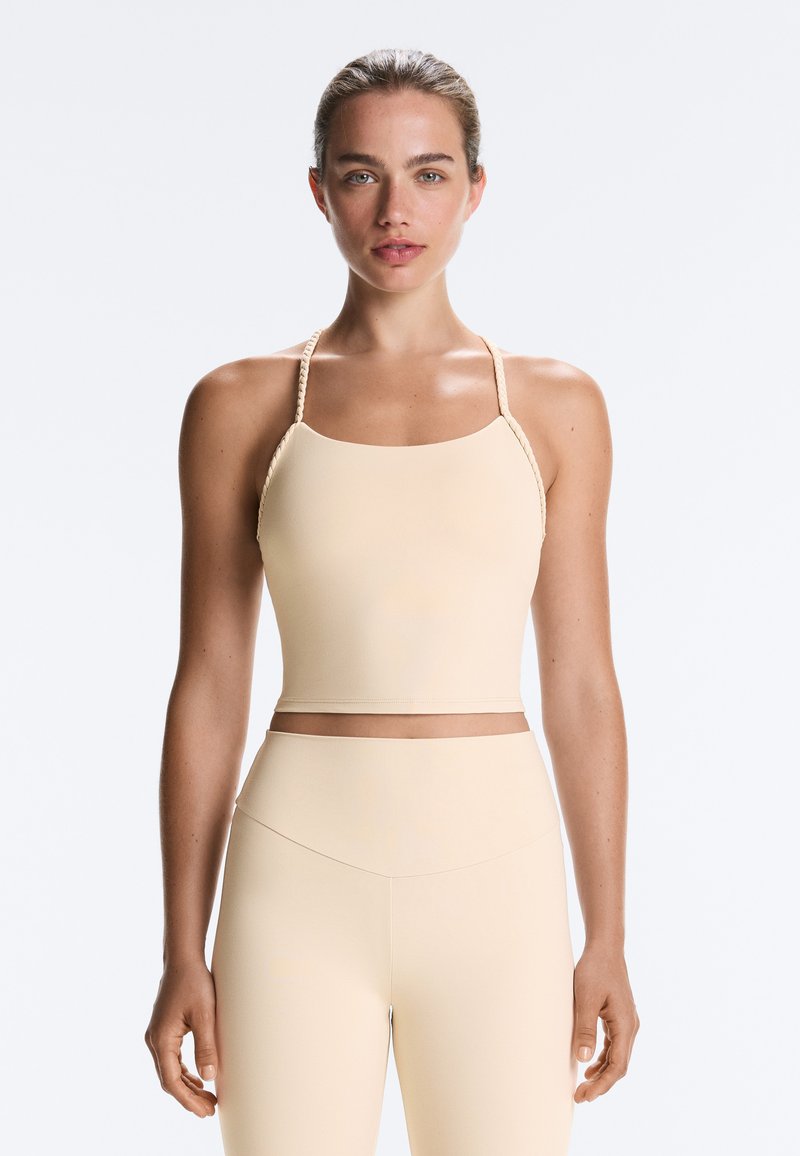 Femme debout, face à l'avant, portant un crop top beige sans manches avec des bretelles fines tressées et un pantalon taille haute assorti.