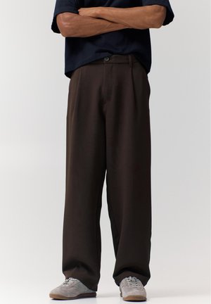 Bărbat purtând pantaloni largi maro închis și adidași gri deschis, stând cu brațele încrucișate pe un fundal simplu, deschis.