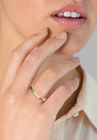 Guld ring med et tekstureret bånd, prydet med flerfarvede ædelsten—grønne, blå, pink og gule—placeret i en lineær rækkefølge.