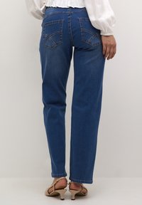 Jeans in denim blu con gamba dritta, caratterizzati da due tasche posteriori e dettagli di cucitura sottili, abbinati a tacchi color nude.