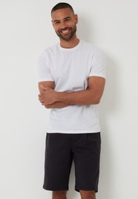 T-shirt en coton blanc à col rond, associé à un short noir jusqu'aux genoux. Le modèle se tient avec les bras croisés devant un fond neutre.