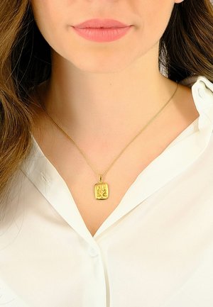 Collier avec pendentif en or, orné d'un charme rectangulaire présentant un design en relief, suspendu à une fine chaîne en or, porté sur un chemisier blanc.