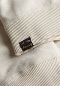 Sudadera color crema con puños acanalados, que presenta una etiqueta azul con la bandera del Reino Unido y la bandera americana bordadas, así como el texto "Superdry".