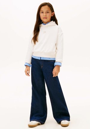 Junges Mädchen steht, trägt einen weißen Hoodie über einem blau gestreiften Hemd, weite dunkle Jeans und weiße Sneakers, vor einem einfarbigen Hintergrund.