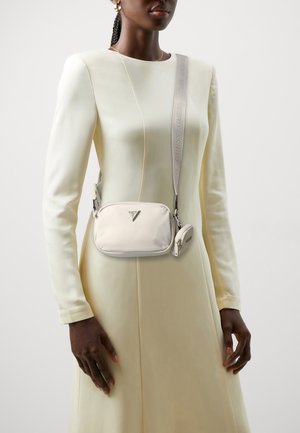 Woman wearing a cream long-sleeve dress and a small beige crossbody bag with a detachable coin pouch and branded strap.