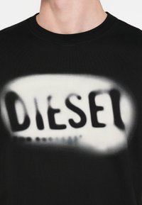 Μαύρο βαμβακερό T-shirt με θολωμένο λευκό λογότυπο σε στυλ ψεκασμού, που γράφει "DIESEL" στο στήθος με λεία υφή.