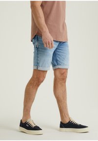 Man som bär uppvikta ljusblå denimshorts, brunt t-shirt och svarta sneakers med vita sulor, som står mot en enfärgad bakgrund.