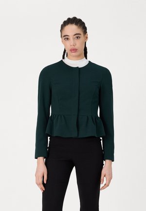 Peplum jacket in dark green fabric, featuring long sleeves, round neckline, and a lined design. Worn over a white collar.