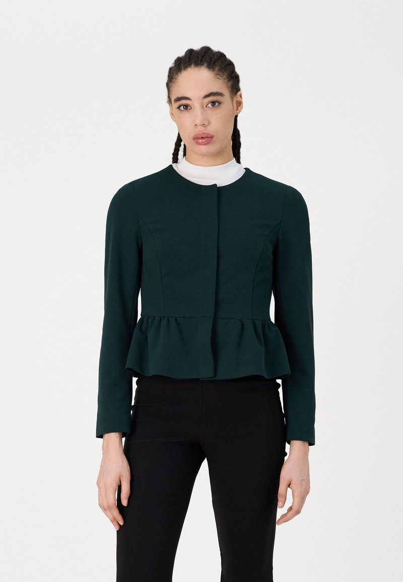Peplum jacket in dark green fabric, featuring long sleeves, round neckline, and a lined design. Worn over a white collar.