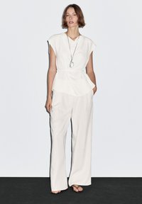 Witte, gedrapeerde blouse met capmouwen en peplum ontwerp, gecombineerd met losse, crèmekleurige wijdbenige broek. Hanger ketting voegt detail toe.