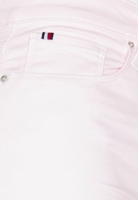 Jeans de mezclilla rosa claro con herrajes plateados, que presentan un pequeño logo multicolor en el bolsillo trasero. Textura suave con un ligero estiramiento.
