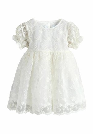 Robe de bébé blanche avec manches courtes bouffantes et superposition de dentelle florale couvrant la robe et ourlet festonné.