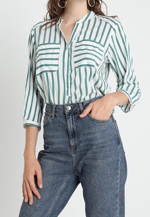 Blusa a righe verdi e bianche con maniche lunghe, due tasche sul petto e colletto, abbinata a jeans in denim blu a vita alta.