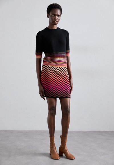 Vestido de malha com parte superior preta e saia em padrão de chevron multicolorido. A textura é suave, com cores vibrantes em degradê que vão do vermelho ao rosa.