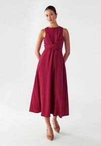 Abito senza maniche color bordeaux con dettaglio anteriore attorcigliato, gonna fluida e lunghezza midi. Tessuto liscio con una consistenza morbida.
