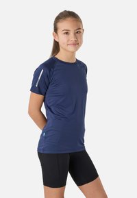 Adolescente debout, les mains derrière le dos, portant un t-shirt de sport bleu marine à manches courtes et un short de cyclisme noir sur fond blanc.