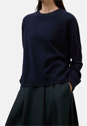 Kvinde iført en mørk marineblå langærmet sweater og en sort plisseret nederdel med hænderne i lommerne, foran en ensfarvet hvid baggrund.