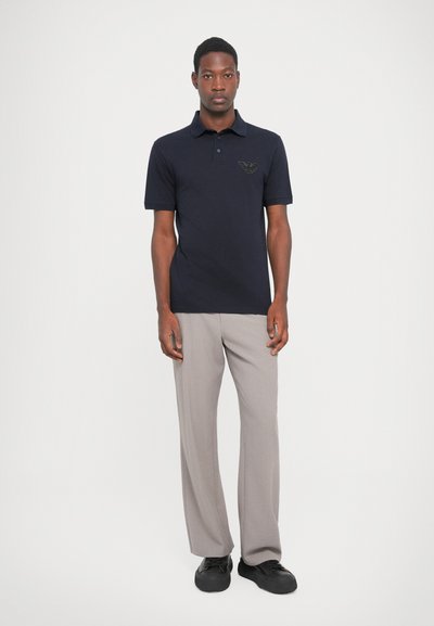 Jūrniecības polo krekls ar apkakli un smalku logotipu, saskaņots ar gaiši pelēkiem biksēm un melnām kurpēm, ar brīvu piegriezumu un gludu tekstūru.