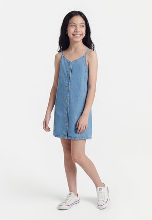 Mosolygó lány, ujjatlan, gombos farmerruhát és fehér tornacipőt visel, egyik lábát kissé előrelépve áll.