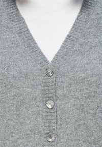 Grau gestrickte Strickjacke mit V-Ausschnitt, ausgestattet mit vier silbernen, strukturierten Knöpfen, über einem weißen Rollkragenoberteil getragen.