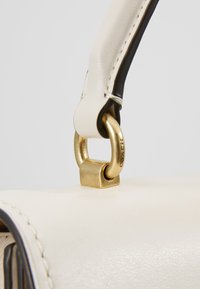 Vitt läderhandtagsrem med guld detaljer, med en rektangulär metallanslutning och svart kant. Slät textur och rena linjer.