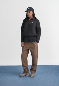 Svart fleecegenser med kvart glidelås, svart caps, brune cargo-bukser og grå sneakers. Jevn tekstur med logo detaljer.