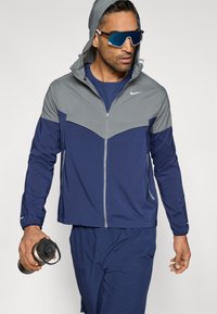 Μπλε σκούρο και γκρι αθλητικό μπουφάν με φερμουάρ και κουκούλα, με σχέδιο σε σχήμα βέλους, συνδυασμένο με αντίστοιχο σύνολο και γυαλιά ηλίου. Κρατάει ένα μπουκάλι νερό στο χέρι.