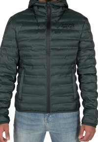 Uomo in giacca a hood in verde scuro, trapuntata, con zip frontale e logo "Wrong Friends", che indossa jeans azzurri.