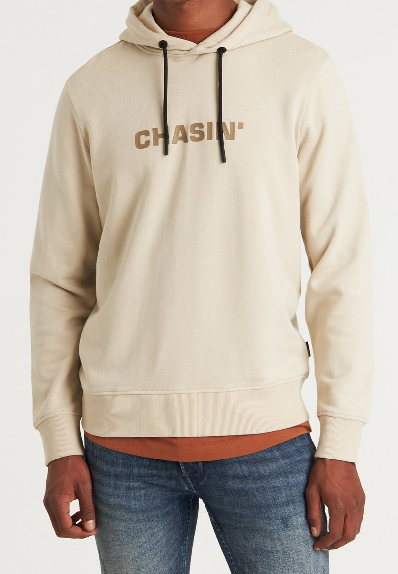 Homme portant un sweat à capuche beige avec le texte « CHASIN' » et des cordons noirs, un t-shirt couleur rouille en dessous, et un jean bleu.