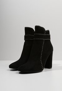 Botas de tobillo de ante negro con una parte superior con forma de onda, tachuelas decorativas a lo largo de la banda y el lado, y un tacón alto y ancho. Textura suave en general.