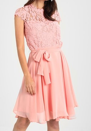 Femme portant une robe rose mi-longue avec un corsage en dentelle, des manches courtes et un nœud à la taille, debout devant un fond uni.