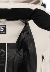 Beige en zwarte jas met een zwart binnenvoering, een ritszak en een label met de tekst "TRAVELIN' waar we lopen" op de bovenrug.