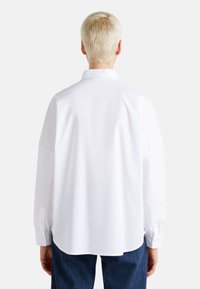 Camicia bianca a maniche lunghe con yoke posteriore, colletto button-down e orlo curvo. Il tessuto appare liscio, con polsini a bottone visibili e una vestibilità morbida.