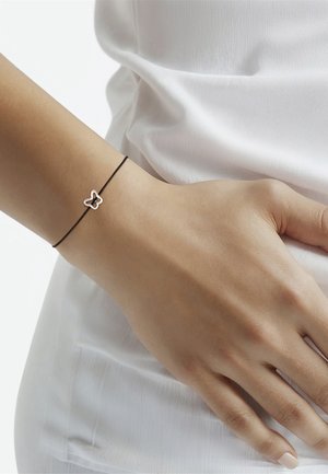Pulsera de cordón negra con un charm de mariposa de plata. El charm está contorneado con un acabado pulido, en contraste con un tono de piel suave.