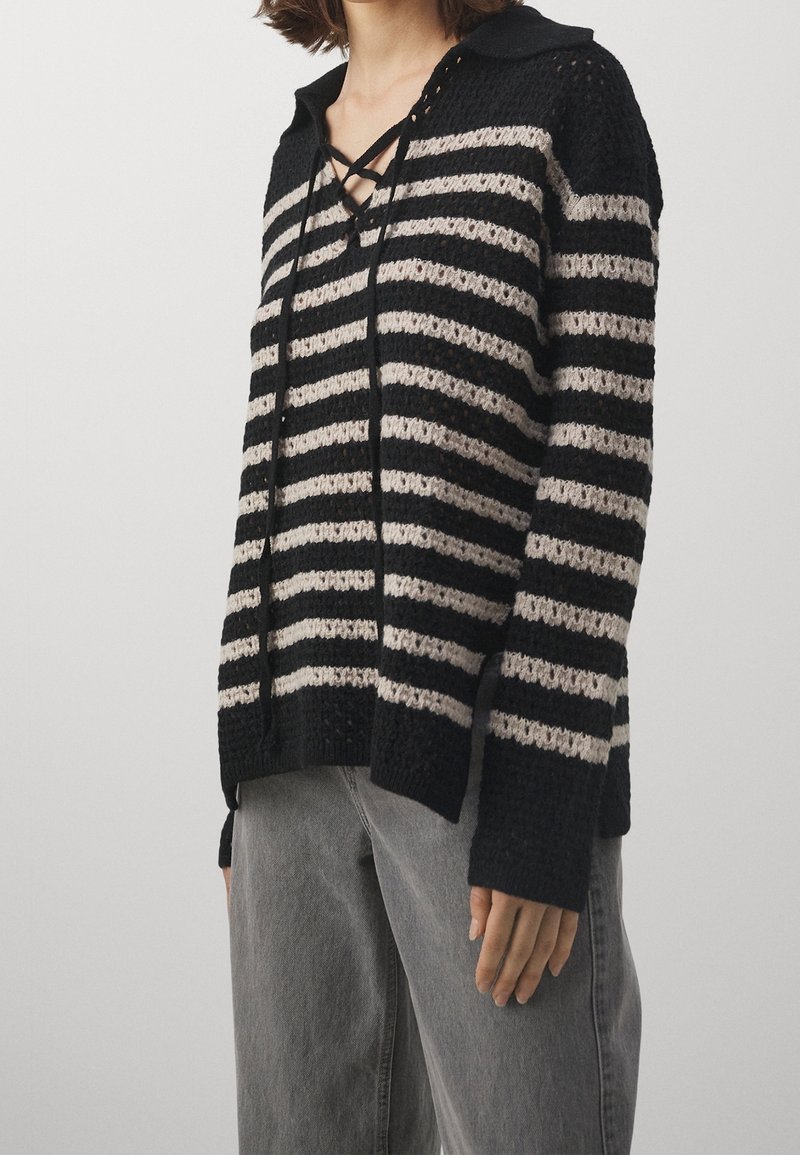 Gestreifter, schwarzer und cremfarbener Strickpullover mit Kragen, Schnürung vorne und lockerer Passform. Der Stoff hat ein strukturiertes Muster und gerippte Bündchen.