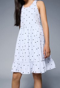 Modré a bílé pruhované šaty s malými černými srdíčky, bez rukávů a s volánkovým lemem. Materiál ze směsi bavlny.