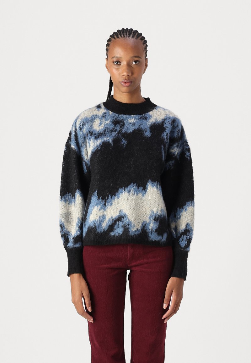 Trui gemaakt van fuzzy breimateriaal met een gedurfde zwarte basis en blauwe en crèmekleurige golfpatronen. Geribbelde halslijn en manchetten. Rode corduroy broek.