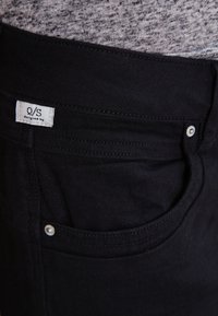 Mustat farkut on valmistettu denimmateriaalista ja niissä on klassinen viiden taskun muotoilu, jossa näkyy brändilogo ja hopeinen nappi taskussa.