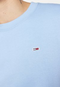 Gros plan sur une chemise bleu clair à col rond montrant un petit logo rectangulaire aux couleurs rouge, blanc et marine sur la poitrine.